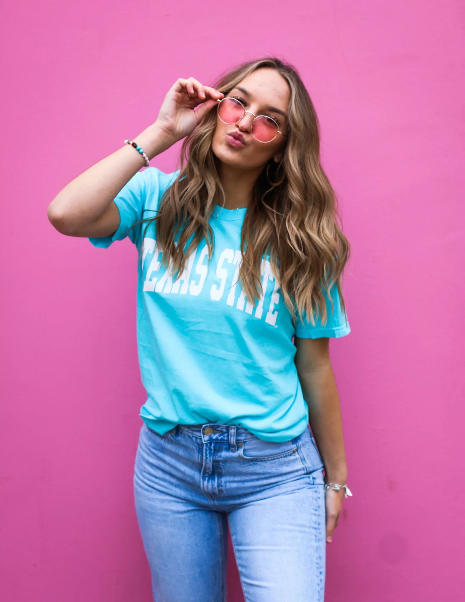 Woman wearing a turquoise 'Texas State' t-shirt and pink sunglasses against a pink background