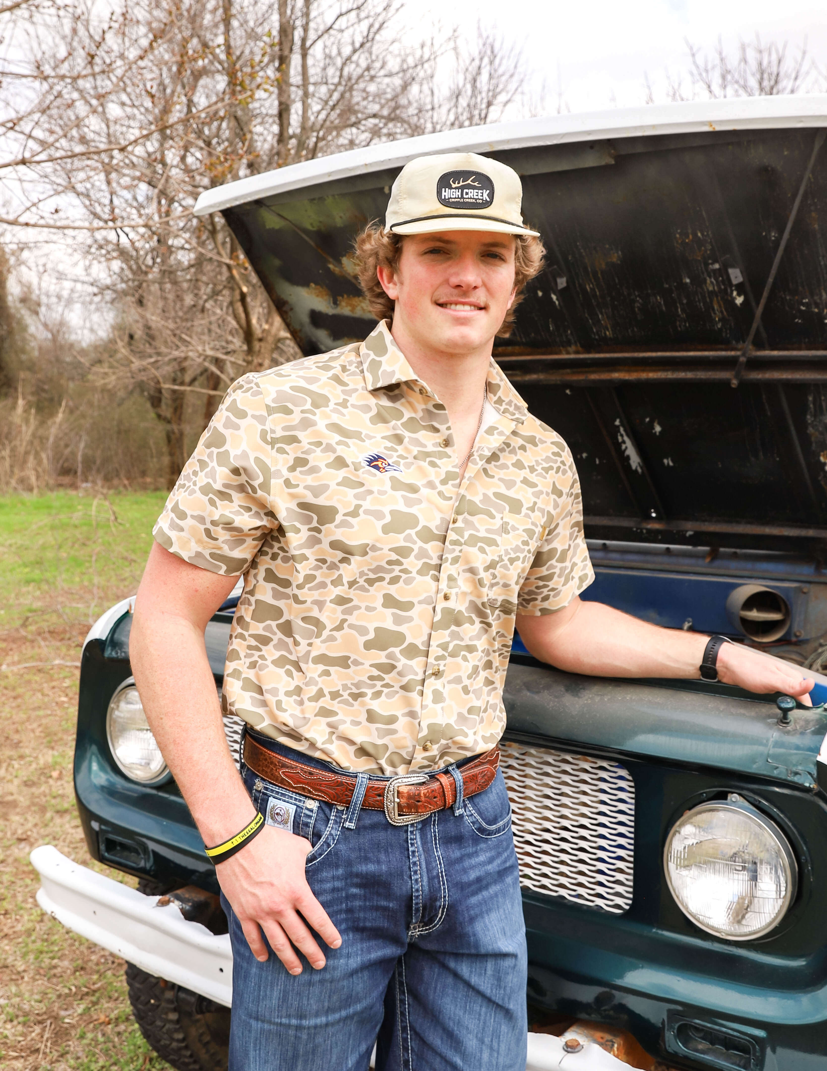 Young man in a camo shirt and cap stands confidently by an open truck hood in a grassy area with bare trees. Casual, outdoorsy vibe.