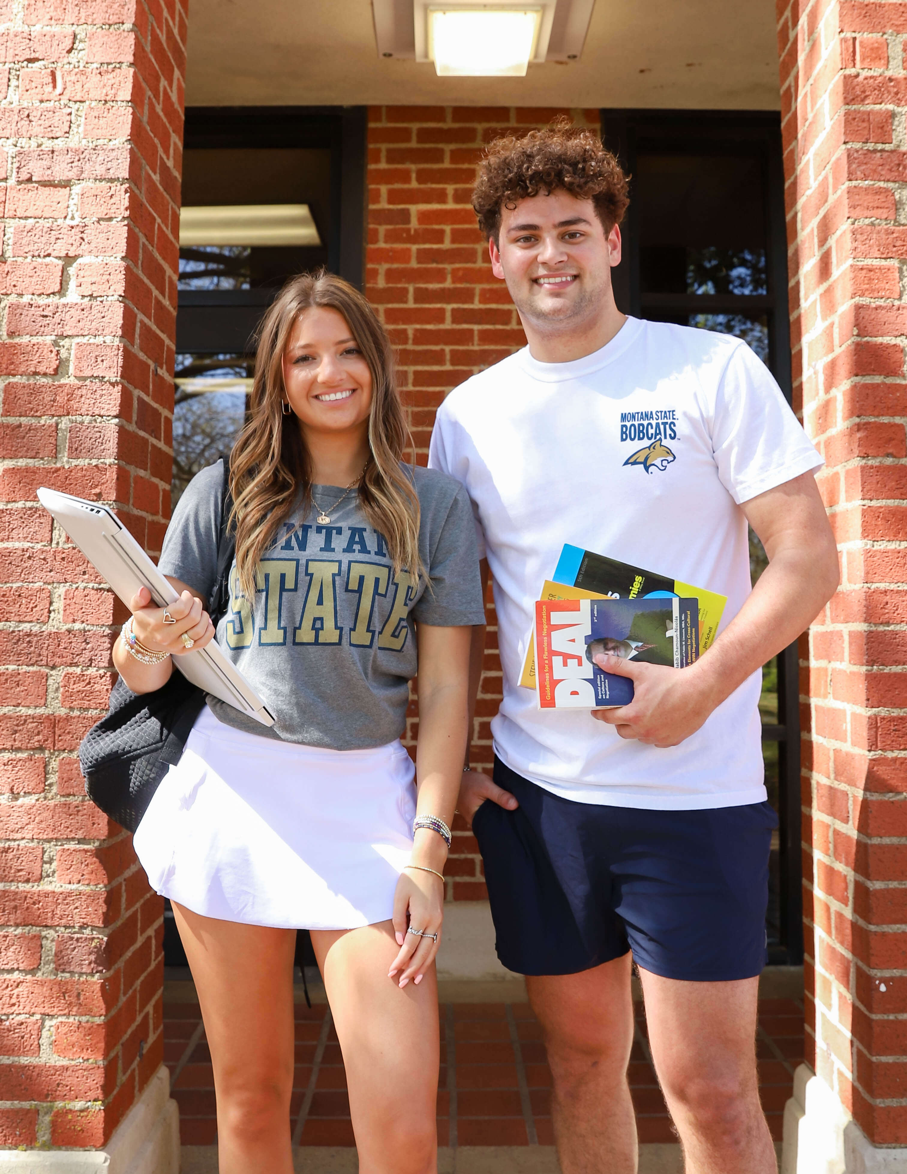Two smiling students stand outside a brick building. One wears a "Montana State" T-shirt, the other holds books. Bright, sunny day, casual university vibe.