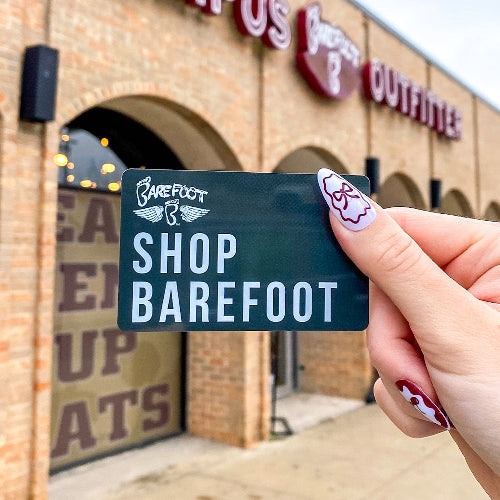 A hand with painted nails holds a "Shop Barefoot" card in front of a brick storefront of Barefoot Campus Outfitter