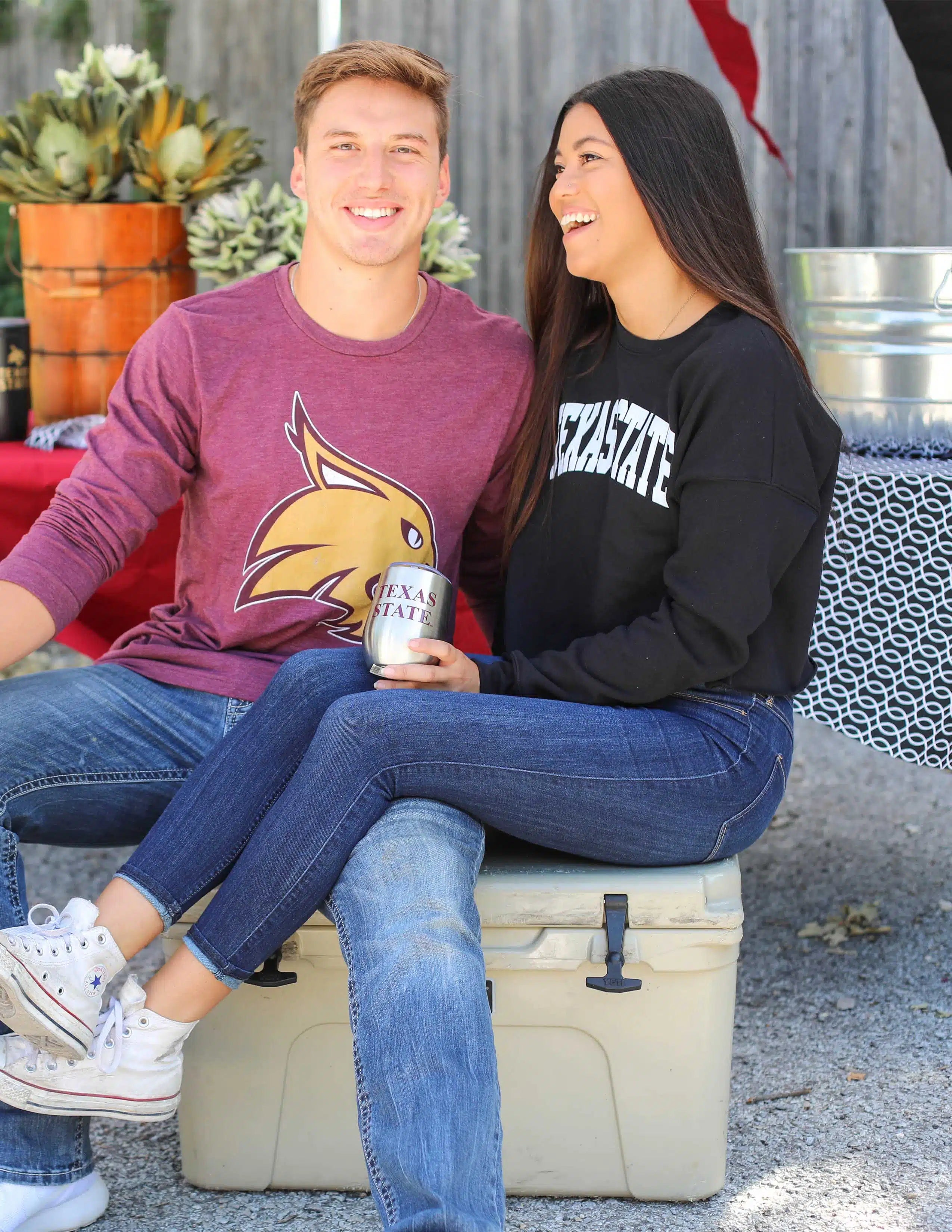 Two people sitting on a cooler outdoors, one holding a can wearing a Texas State Bobcats long sleeve shirt.