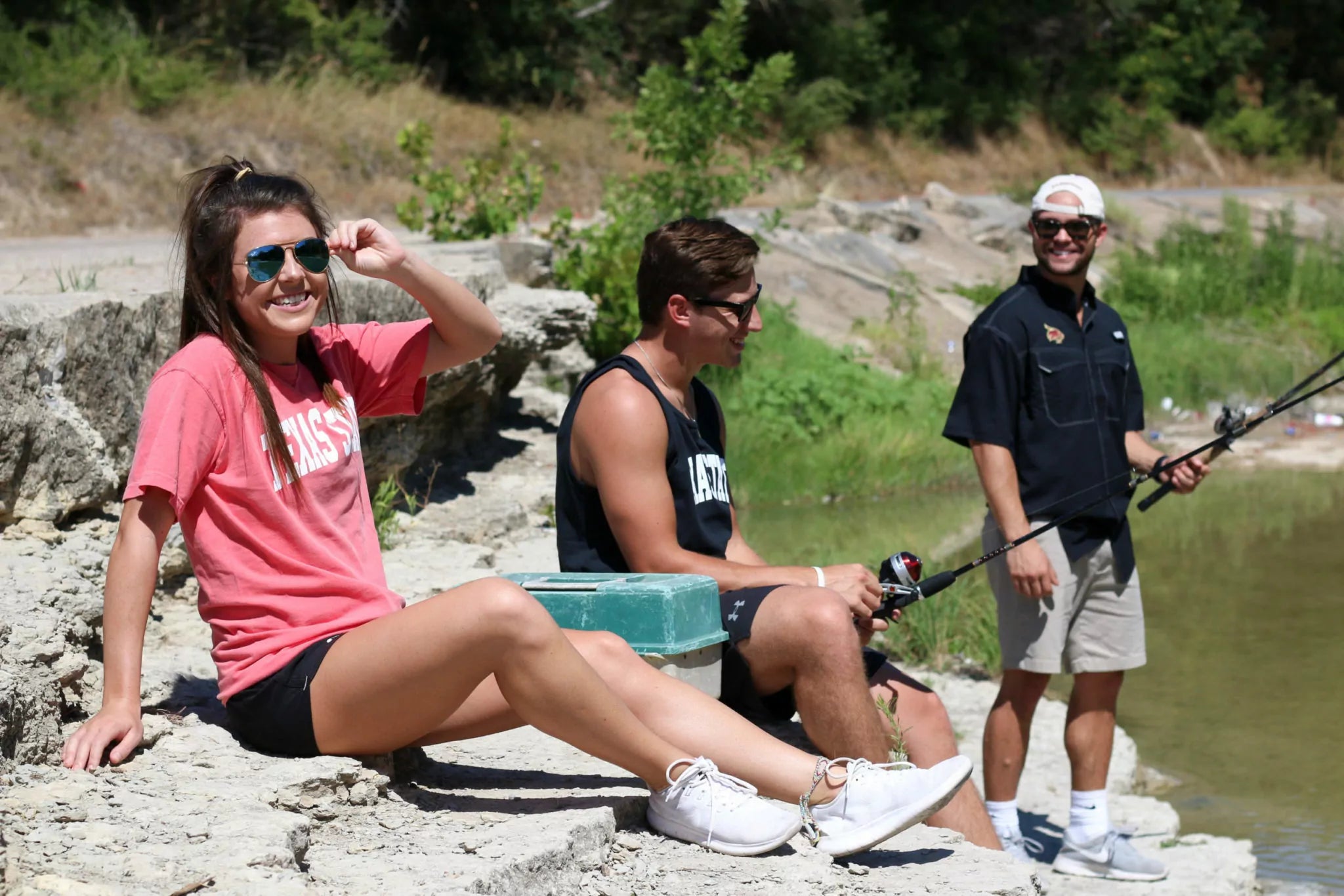 Three people by a lake, two fishing and one sitting on rocks wearing a pink Texas State short sleeve shirt