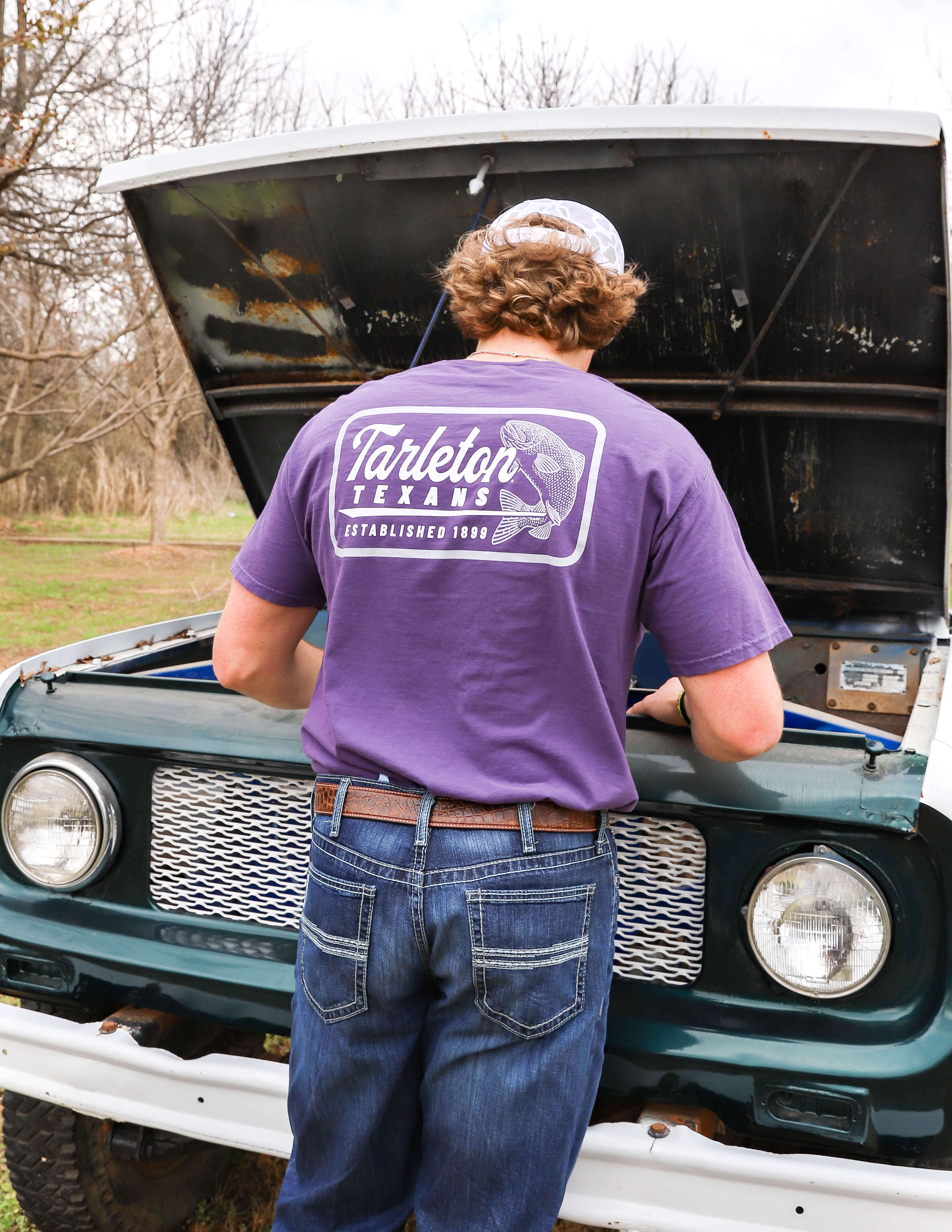 A person in a purple "Tarleton Texans" shirt stands by an open car hood, working on the engine. The setting is outdoors, with bare trees visible.