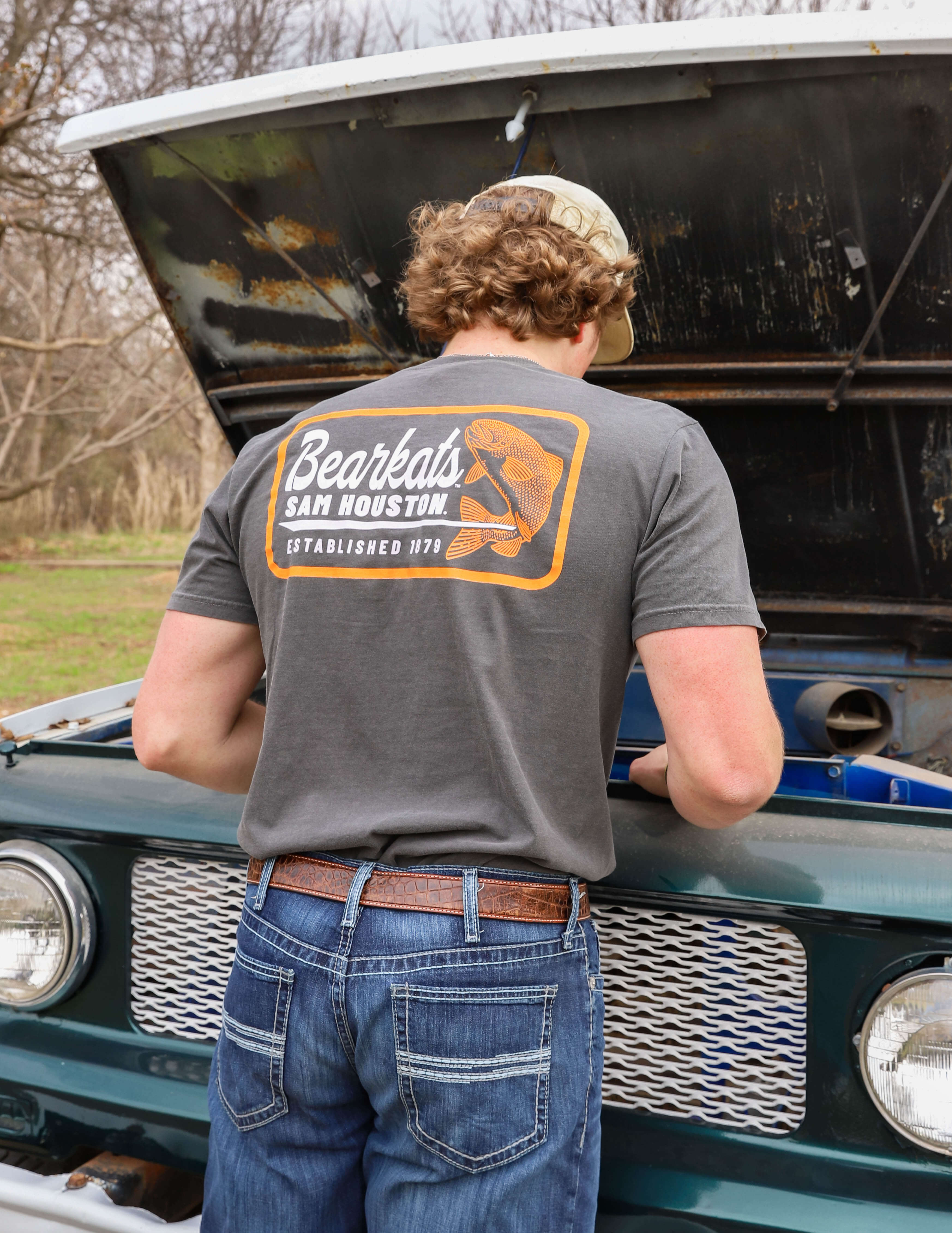 A guy wearing a "Bearkats Sam Houston" T-shirt and jeans works under the hood of an old truck. The scene is outdoors on a cloudy day.