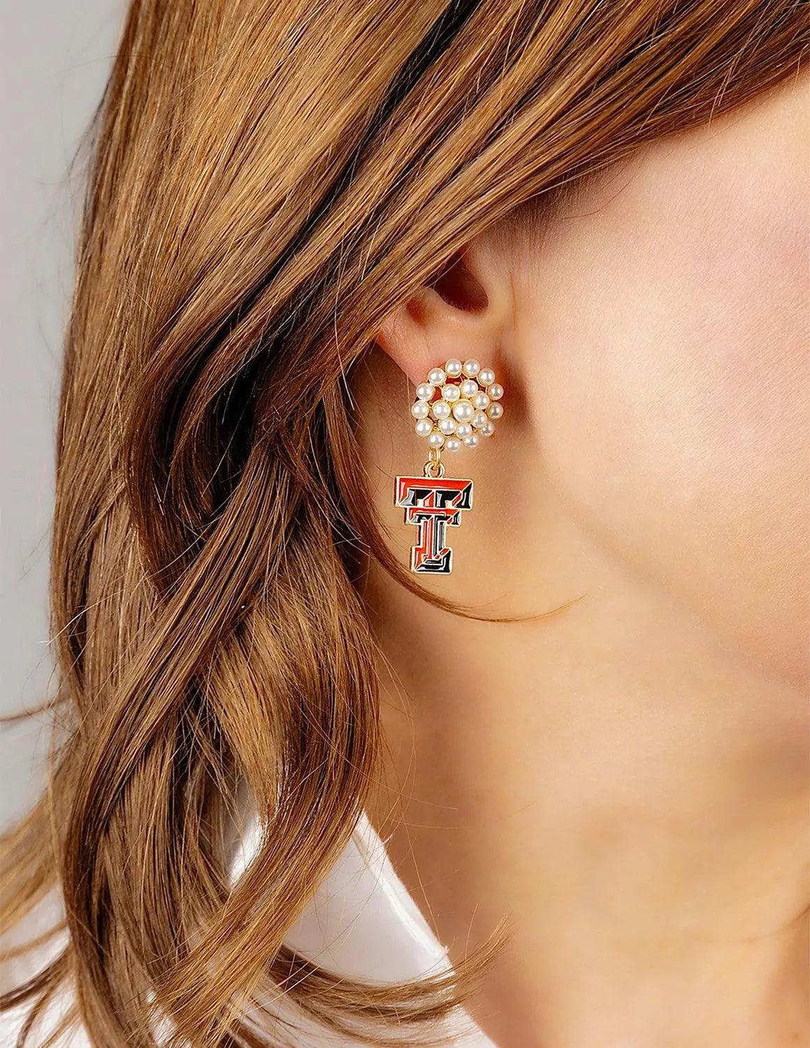 Close-up of a model wearing Texas Tech University earrings with a pearl cluster and a connected double T logo charm underneath.